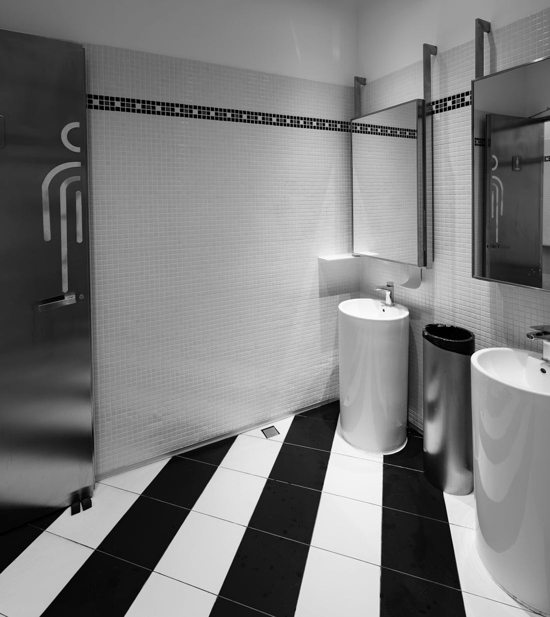 A modern restroom with white tiled walls, pedestal sinks, and black and white striped flooring